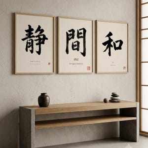 Op de afbeelding: Drie ingelijste wandkunstwerken met zwarte Japanse kalligrafie op een gebroken witte achtergrond. De prints tonen de woorden SHIZUKA, ma en wa met hun Engelse vertalingen eronder. Onder de kunstwerken staat een lange houten consoletafel.