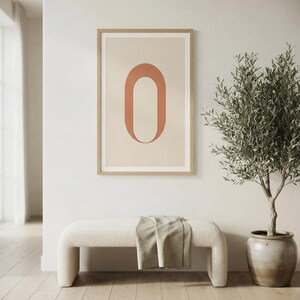 Op de afbeelding: Een ingelijste kunstprint met een grote, terracotta-kleurige letter O gecentreerd op een lichtbeige achtergrond. Het kunstwerk zit in een dunne, lichtgekleurde houten lijst en toont een strak, grafisch ontwerp met zachte, neutrale tinten.