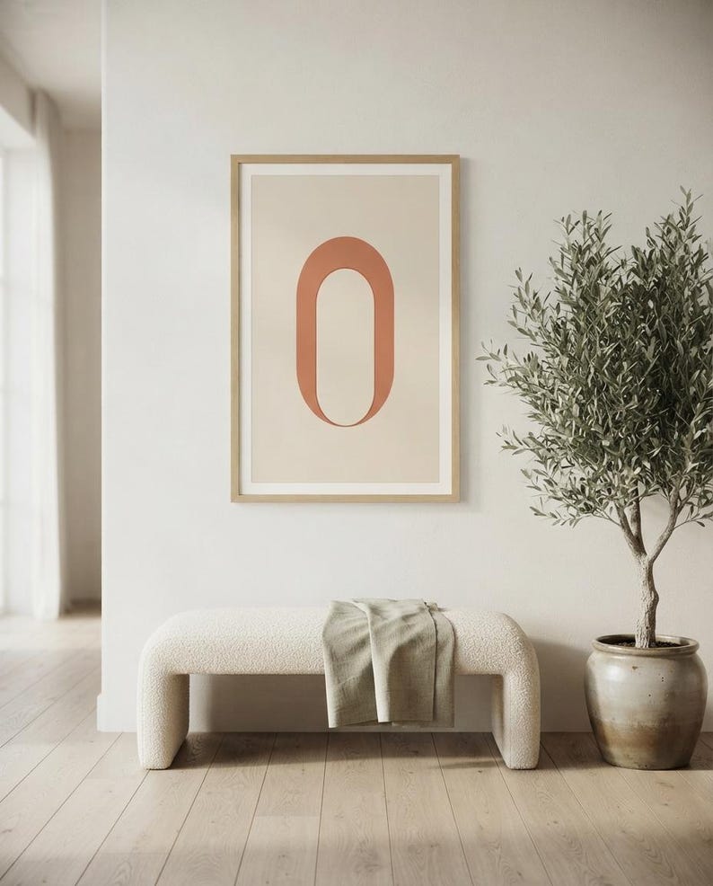 Op de afbeelding: Een ingelijste kunstprint met een grote, terracotta-kleurige letter O gecentreerd op een lichtbeige achtergrond. Het kunstwerk zit in een dunne, lichtgekleurde houten lijst en toont een strak, grafisch ontwerp met zachte, neutrale tinten.
