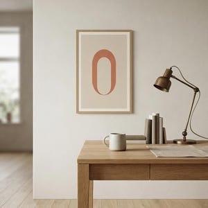 Op de afbeelding: Een ingelijste wandkunstprint met een grote, terracotta-kleurige letter O gecentreerd op een lichtbeige achtergrond. De print zit in een eenvoudige, lichtgekleurde houten lijst en hangt aan een muur boven een houten bureau.