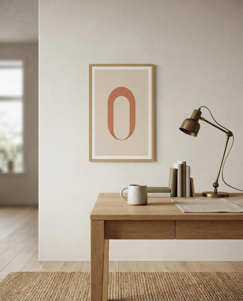 Op de afbeelding: Een ingelijste wandkunstprint met een grote, terracotta-kleurige letter O gecentreerd op een lichtbeige achtergrond. De print zit in een eenvoudige, lichtgekleurde houten lijst en hangt aan een muur boven een houten bureau.