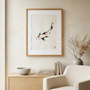 A framed wall art print featuring a sumi-e style ink wash painting of a single koi fish. The fish is white with black markings and orange spots, swimming diagonally. A small red square seal is visible in the bottom right corner.