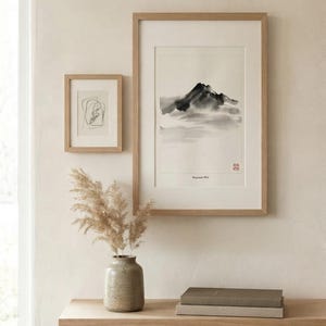 Two framed art prints in light wood frames. The larger vertical print features a black ink wash mountain landscape with the text Mountain Mist and a red seal. The smaller print shows a black line drawing. A ceramic vase holds dried grass.