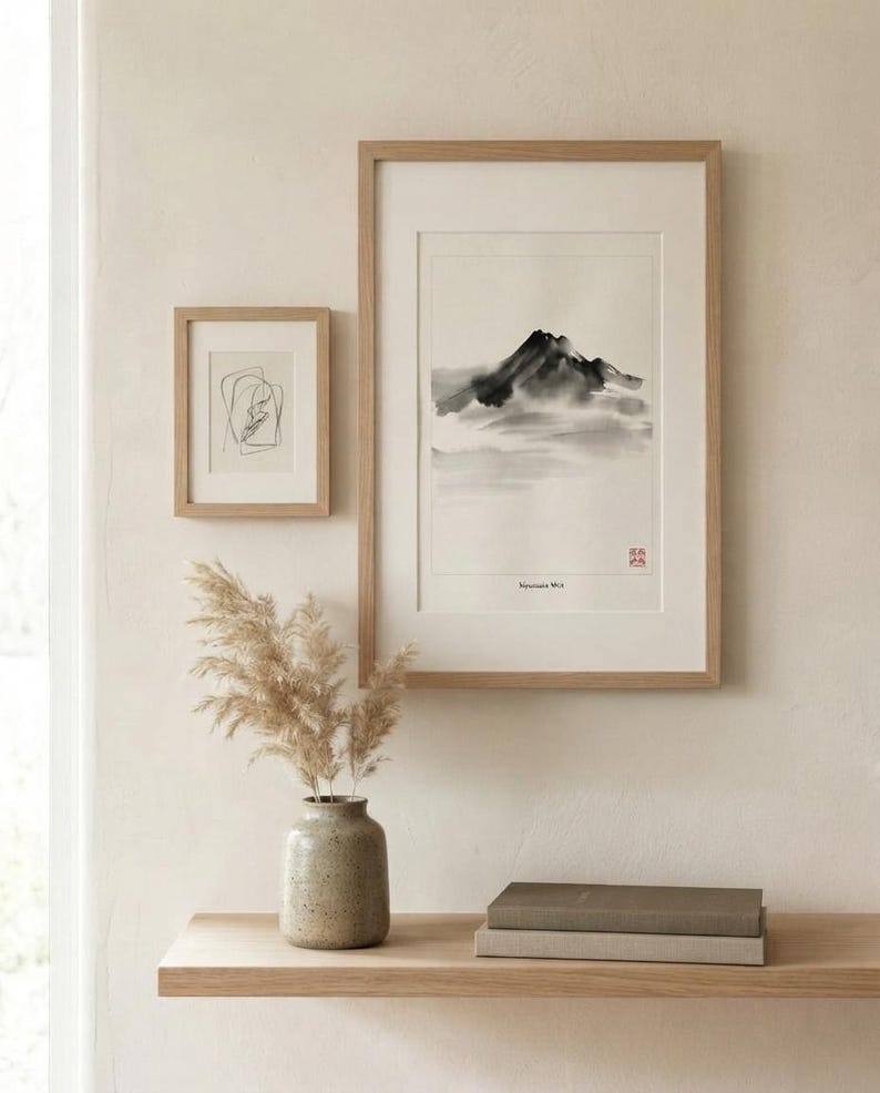 Two framed art prints in light wood frames. The larger vertical print features a black ink wash mountain landscape with the text Mountain Mist and a red seal. The smaller print shows a black line drawing. A ceramic vase holds dried grass.