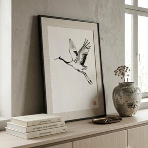 Sumi-e crane print framed in oak, seen on three quiet walls — above a linen bed, on a gallery wall and above an oak console.