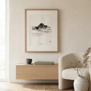 A framed wall art print titled Mountain Mist featuring a black ink wash painting of a mountain peak partially obscured by clouds. The artwork is enclosed in a light-colored wooden frame with a white mat board and a small red seal stamp.