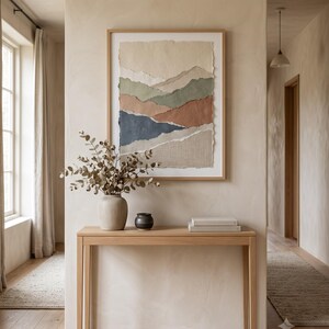 A framed wall art piece featuring layered, torn-edge paper in shades of cream, sage green, terracotta, and navy blue, displayed above a light wood console table holding a ceramic vase with dried eucalyptus and a small black bowl.