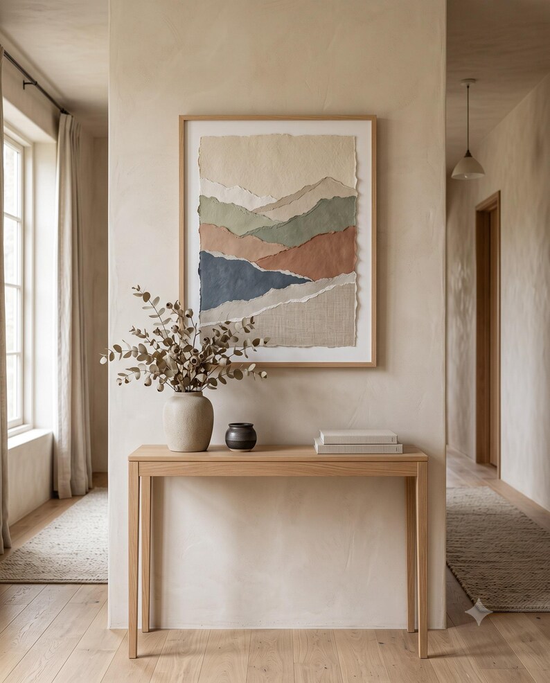 A framed wall art piece featuring layered, torn-edge paper in shades of cream, sage green, terracotta, and navy blue, displayed above a light wood console table holding a ceramic vase with dried eucalyptus and a small black bowl.