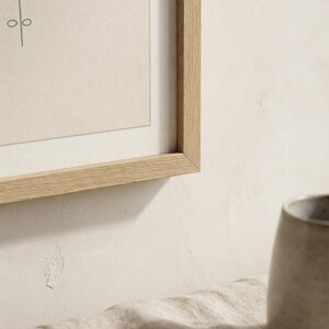 May include: A light oak wood picture frame containing a beige print with a thin dark line drawing of a vertical stem with circular leaves. The frame is positioned above a textured beige fabric and a portion of a grey ceramic mug.