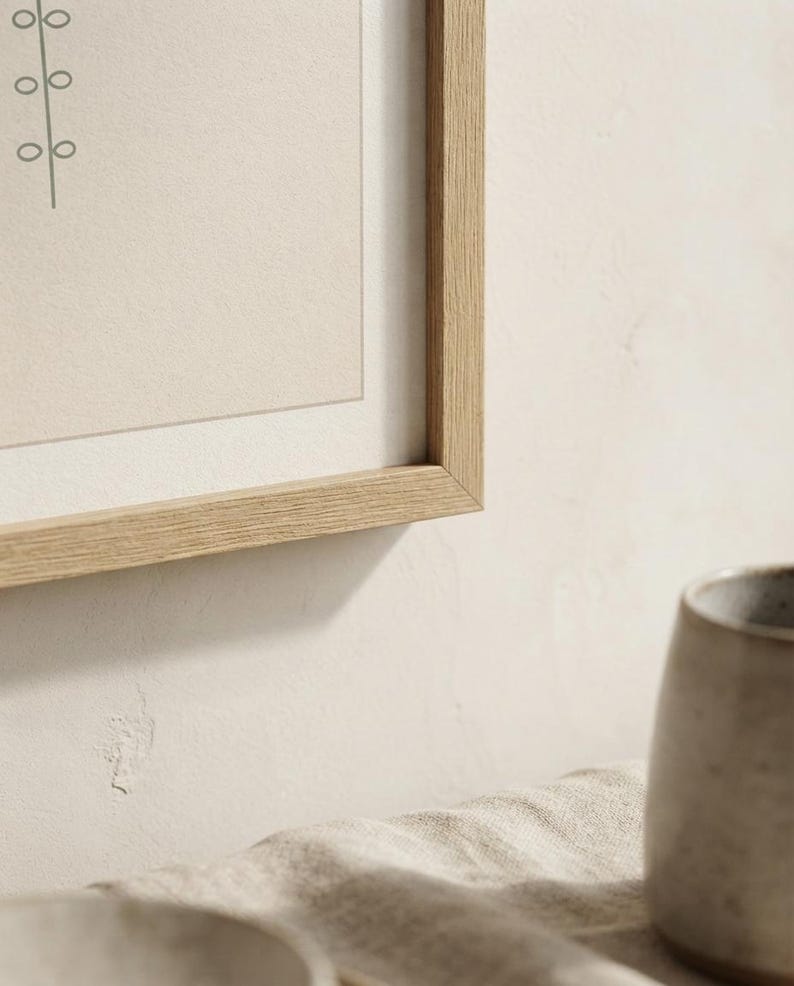 May include: A light oak wood picture frame containing a beige print with a thin dark line drawing of a vertical stem with circular leaves. The frame is positioned above a textured beige fabric and a portion of a grey ceramic mug.