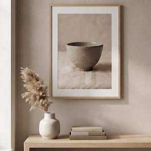 Può includere: Una stampa d'arte incorniciata raffigurante una ciotola in ceramica grigio chiaro riparata con linee kintsugi dorate. L'opera è esposta sopra una console in legno chiaro con un vaso bianco contenente erba della pampa secca e tre libri.