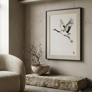Sumi-e crane print framed in oak, seen on three quiet walls — above a linen bed, on a gallery wall and above an oak console.