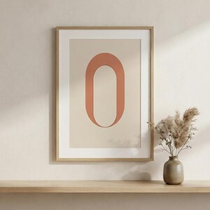 Op de afbeelding: Een ingelijste wandkunstprint met een grote, terracotta-kleurige letter O gecentreerd op een lichtbeige achtergrond. Het kunstwerk zit in een dunne, lichtgekleurde houten lijst en toont een strak, geometrisch ontwerp met een matte afwerking.