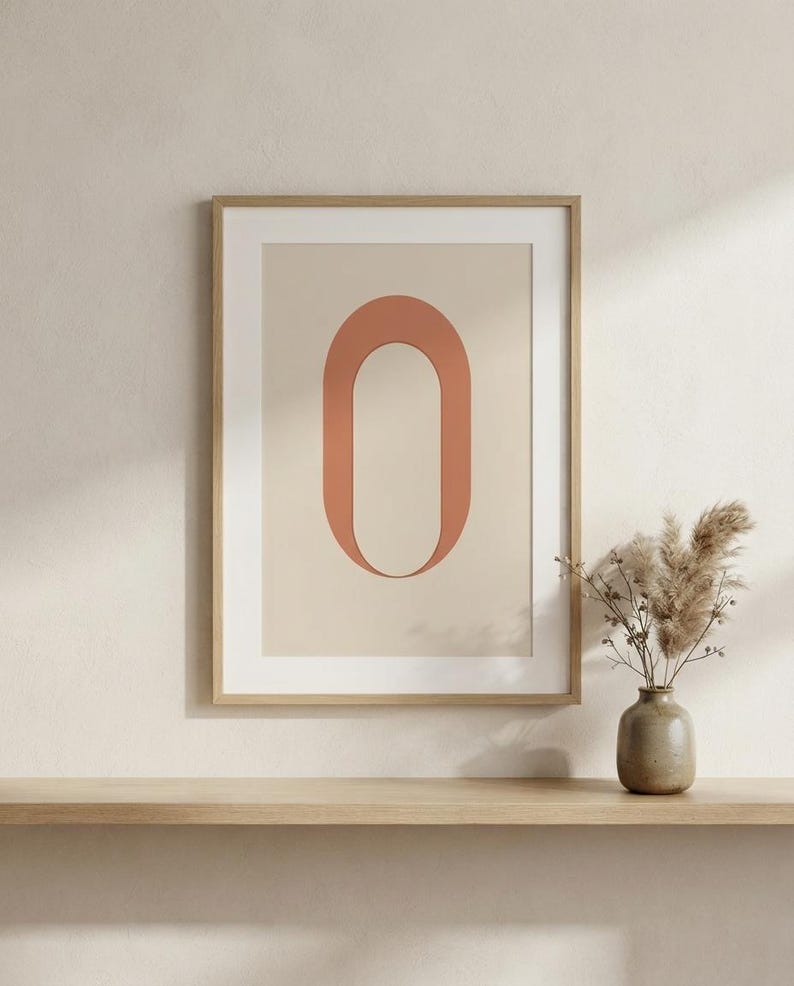 Op de afbeelding: Een ingelijste wandkunstprint met een grote, terracotta-kleurige letter O gecentreerd op een lichtbeige achtergrond. Het kunstwerk zit in een dunne, lichtgekleurde houten lijst en toont een strak, geometrisch ontwerp met een matte afwerking.