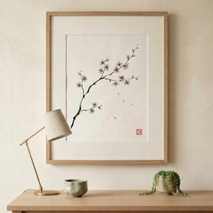 A framed wall art print featuring a black ink wash painting of a flowering cherry blossom branch on a white background. The artwork is enclosed in a light-toned wooden frame and includes a small red square seal stamp in the bottom corner.