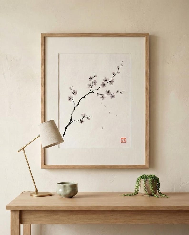 A framed wall art print featuring a black ink wash painting of a flowering cherry blossom branch on a white background. The artwork is enclosed in a light-toned wooden frame and includes a small red square seal stamp in the bottom corner.