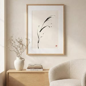 A framed wall art print featuring a black ink brush stroke illustration of a single blade of grass on a cream background. The artwork is enclosed in a light wood frame with a white mat and includes a small red square stamp in the corner.