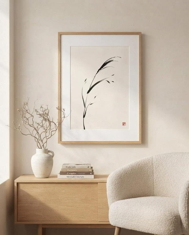 A framed wall art print featuring a black ink brush stroke illustration of a single blade of grass on a cream background. The artwork is enclosed in a light wood frame with a white mat and includes a small red square stamp in the corner.