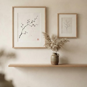 Two framed wall art prints with light wood frames. The larger print features a black ink drawing of a flowering branch with a red stamp. The smaller print shows a delicate line drawing of bare twigs. A ceramic vase holds dried pampas grass.