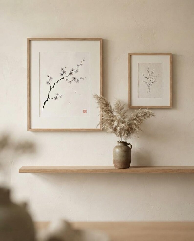 Two framed wall art prints with light wood frames. The larger print features a black ink drawing of a flowering branch with a red stamp. The smaller print shows a delicate line drawing of bare twigs. A ceramic vase holds dried pampas grass.