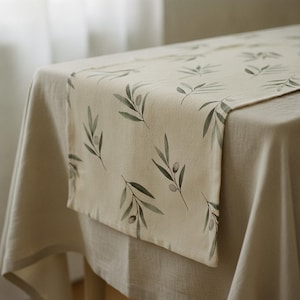 May include: A rectangular table runner featuring a repeating pattern of green olive branches and small olives printed on a light beige fabric. The runner is draped over a solid light beige tablecloth, showcasing a smooth, woven textile texture.