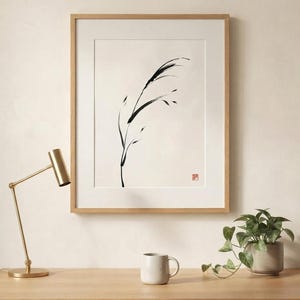 A framed wall art print featuring a black ink brush stroke illustration of a single blade of grass on a cream background. The artwork is enclosed in a light wood frame and includes a small red square stamp in the bottom right corner.