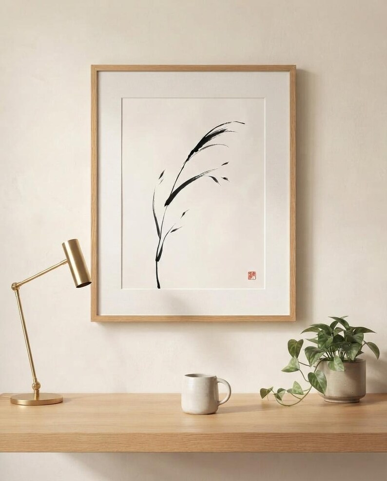 A framed wall art print featuring a black ink brush stroke illustration of a single blade of grass on a cream background. The artwork is enclosed in a light wood frame and includes a small red square stamp in the bottom right corner.
