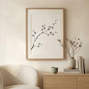 A framed wall art print featuring a black ink illustration of a delicate cherry blossom branch with small light pink flowers on a cream background, encased in a light-toned wooden frame with a white mat border.