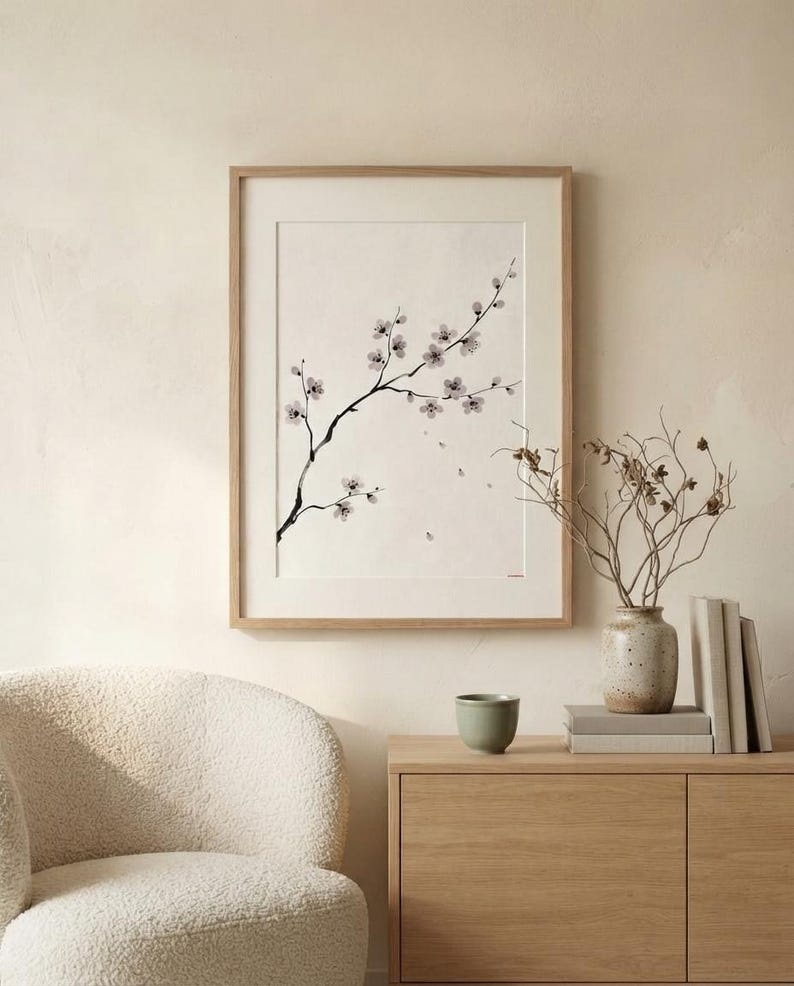 A framed wall art print featuring a black ink illustration of a delicate cherry blossom branch with small light pink flowers on a cream background, encased in a light-toned wooden frame with a white mat border.