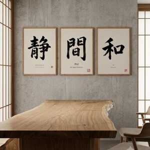 Op de afbeelding: Een set van drie ingelijste wandkunstprints met zwarte Japanse kalligrafie op een cr&egrave;mekleurige achtergrond. Elke print bevat Engelse tekst onder de tekens met de woorden SHIZUKA, ma en wa, samen met een klein rood stempelontwerp.