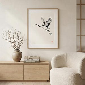 A framed wall art print featuring a black and white ink illustration of a crane in flight on a cream background. The artwork includes a small red square seal in the bottom right corner and is housed in a light wood frame.