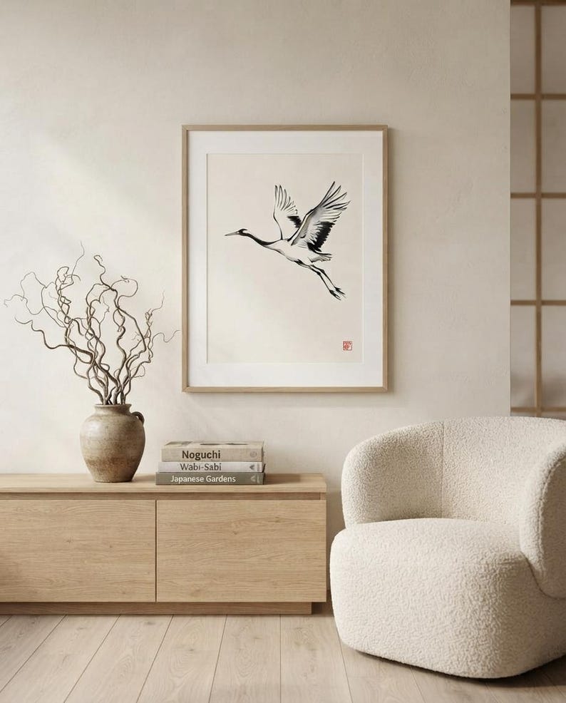 A framed wall art print featuring a black and white ink illustration of a crane in flight on a cream background. The artwork includes a small red square seal in the bottom right corner and is housed in a light wood frame.