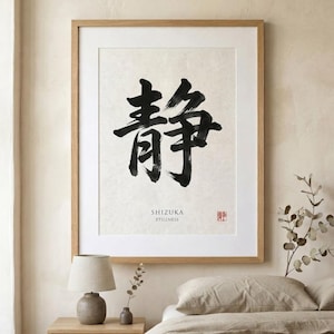 A framed wall art print featuring a large black ink brush calligraphy character for stillness on a textured off-white background. Below the character, the text reads SHIZUKA STILLNESS, accompanied by a small red square seal stamp.