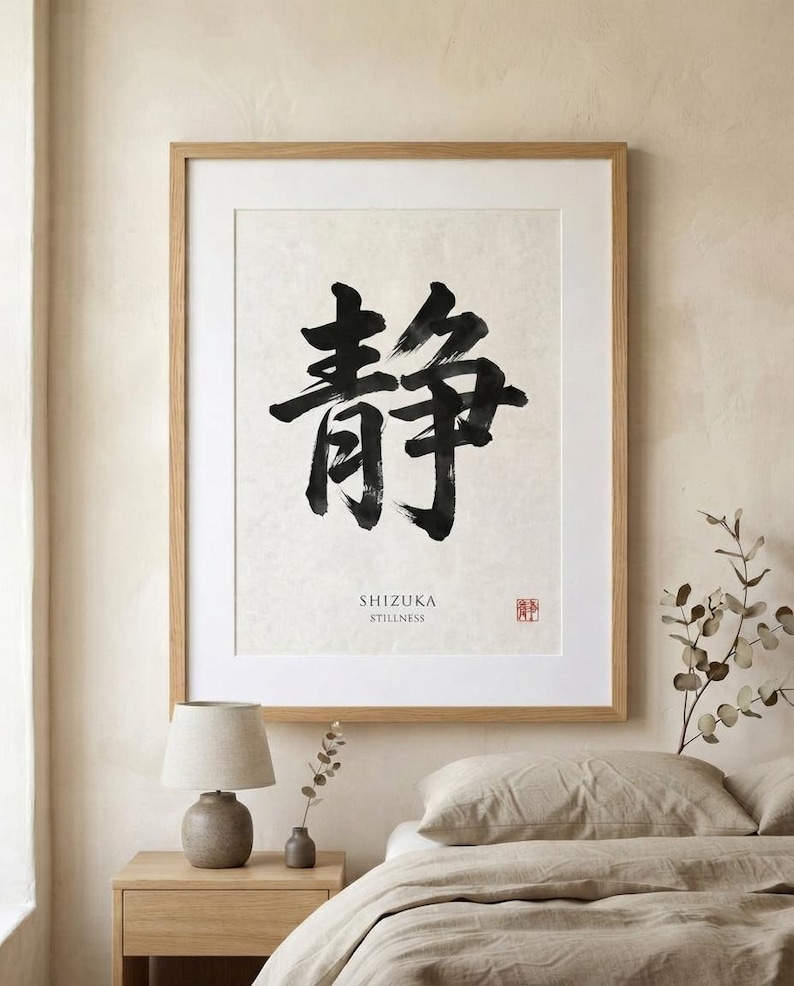 A framed wall art print featuring a large black ink brush calligraphy character for stillness on a textured off-white background. Below the character, the text reads SHIZUKA STILLNESS, accompanied by a small red square seal stamp.