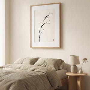 A framed wall art print featuring a black ink brush stroke illustration of a single blade of grass on a cream background, displayed in a light wood frame with a white mat, hanging above a bed with beige linen bedding.