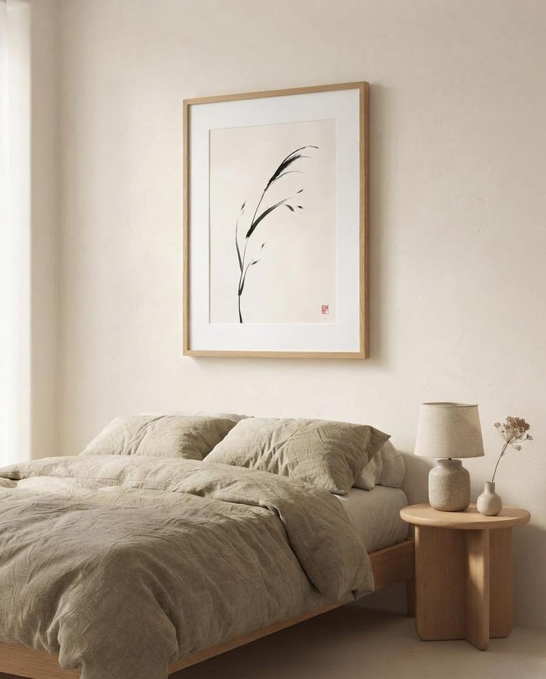 A framed wall art print featuring a black ink brush stroke illustration of a single blade of grass on a cream background, displayed in a light wood frame with a white mat, hanging above a bed with beige linen bedding.