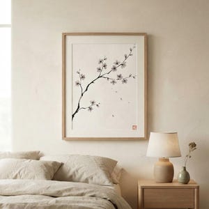 A framed wall art print featuring a black ink wash painting of a delicate cherry blossom branch with small white flowers and buds on a cream background, enclosed in a light-toned wooden frame with a small red square seal in the corner.