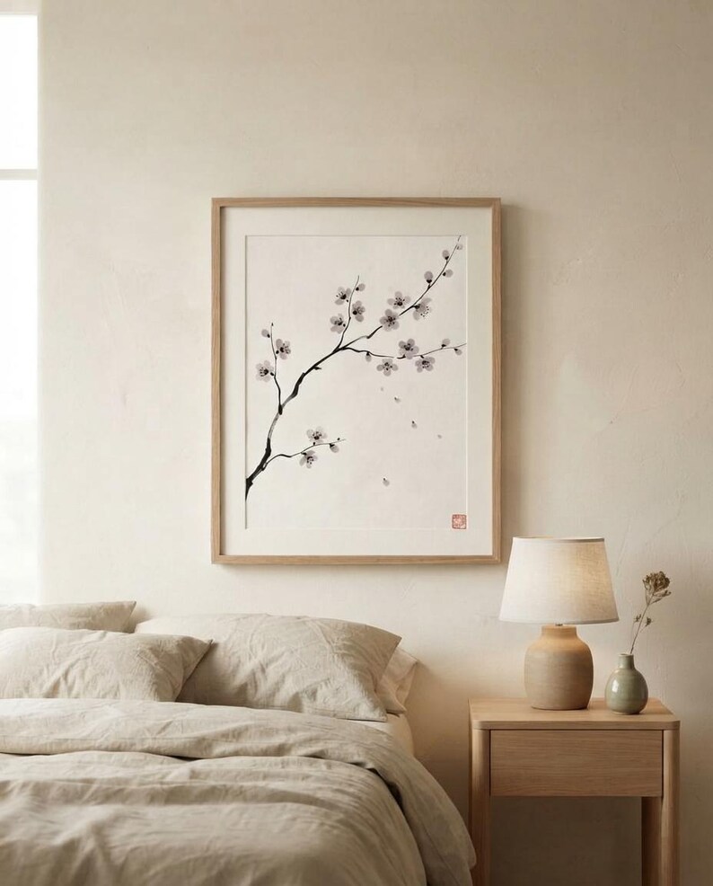 A framed wall art print featuring a black ink wash painting of a delicate cherry blossom branch with small white flowers and buds on a cream background, enclosed in a light-toned wooden frame with a small red square seal in the corner.