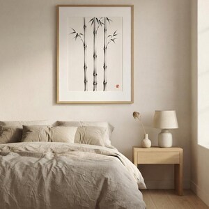 A framed wall art print featuring a black and white ink wash painting of three bamboo stalks. The artwork is displayed in a light wood frame with a white mat border, showcasing detailed nodes and leaves with a small red seal stamp.