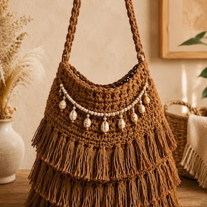 May include: A brown crocheted shoulder bag featuring three horizontal tiers of long fringe along the bottom. The bag includes a braided strap and a decorative strand of small white beads and off-white seashells draped across the front.