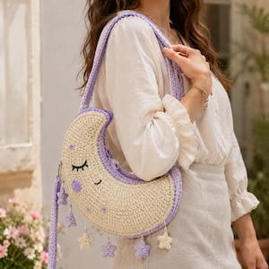 May include: A crescent moon-shaped crochet shoulder bag in cream and lavender. The bag features a sleeping face with embroidered eyes and mouth, small purple star accents, and dangling star-shaped charms along the bottom edge with a strap.