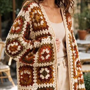 May include: A long-sleeved cardigan made of crocheted granny squares in a color palette of cream, rust orange, mustard yellow, and olive green. The garment features an open front design with a cream-colored border along the edges and sleeves.