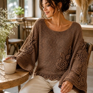 May include: A brown long-sleeved sweater featuring a wide scoop neckline and flared sleeves. The garment is decorated with three-dimensional crocheted floral and leaf appliques on the sleeves and front, finished with a scalloped hemline.