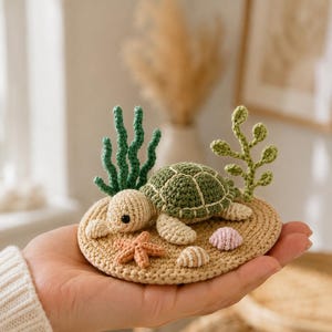 Puede incluir: Una pequeña base redonda de ganchillo beige con una tortuga de color beige claro y caparazón verde oscuro. La escena incluye una rama de algas verde oscuro, una planta de hojas verde claro, una pequeña estrella de mar naranja y dos conchas.