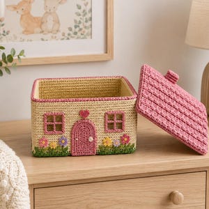 May include: A rectangular crochet storage basket designed to look like a small house with a cream-colored body, a pink arched door, two pink-framed windows, and a removable pink roof lid. The base features colorful crocheted flowers and green grass.