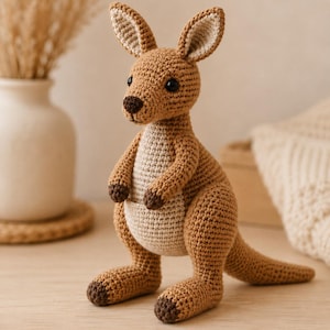 May include: A crocheted kangaroo toy made with brown yarn, featuring a lighter beige belly pouch, dark brown yarn accents on the paws and nose, and two upright ears. The stuffed animal has black plastic eyes and a sturdy tail for balance.