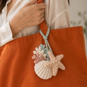 Puede incluir: Un bolso tote grande de color naranja con un accesorio de ganchillo colgante. El accesorio consta de un cordón azul claro unido a una concha blanca, una estrella de mar beige con cuentas blancas y dos pequeñas piezas con forma de coral en rosa y azul.