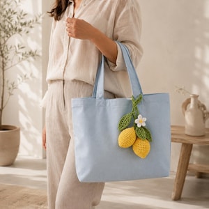 May include: A light blue canvas tote bag featuring a decorative crochet accessory attached to the handle. The accessory consists of two yellow lemons, green leaves, and a small white flower with a yellow centre, all crafted from yarn.