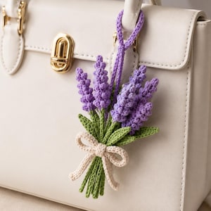 May include: A crochet lavender flower bouquet bag charm featuring purple flower heads, green stems and leaves, and a beige bow, attached to a cream-colored handbag with gold hardware. The accessory is crafted with detailed yarn stitching.