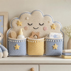 May include: A cream-colored crochet wall organizer shaped like a cloud with a smiling face, featuring three pockets in light blue and yellow. The pockets are decorated with white faux pearls and yellow crochet stars, holding a bottle and cloth.